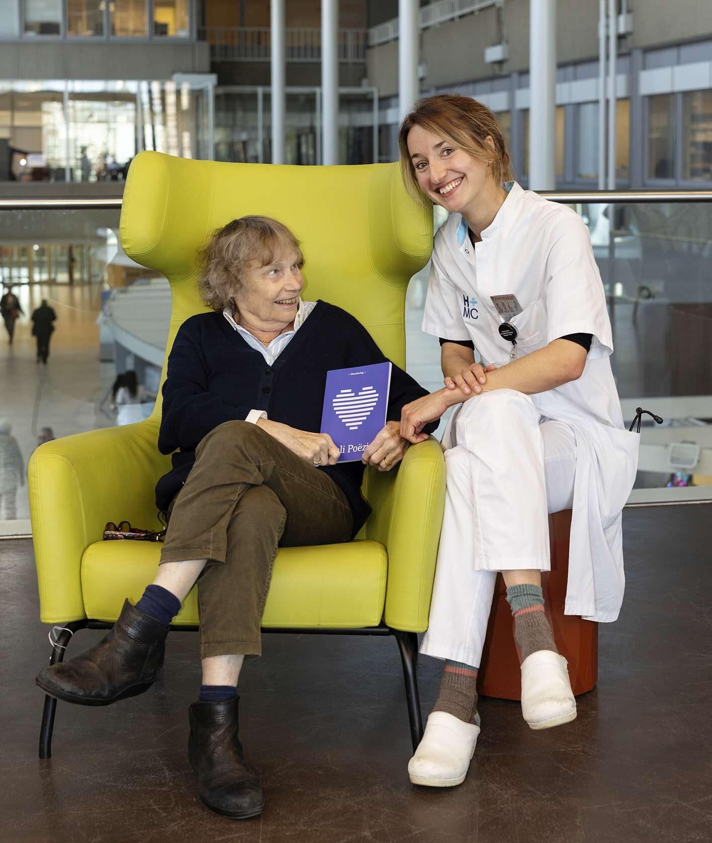 Basisarts Julia met een patiënt op de foto. De patiënt zit in de stoel en samen houden ze een dichtbundel vast, speciaal gemaakt voor de afdeling cardiologie